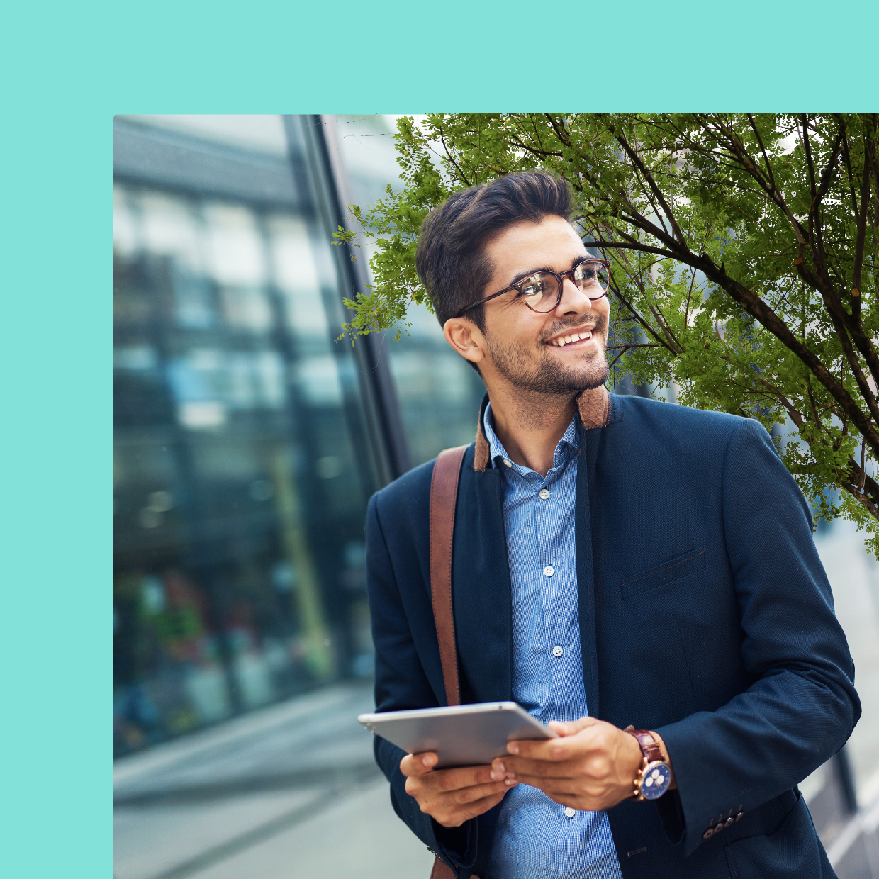 A young businessman in a dark blazer and glasses holding a tablet and looking thoughtfully upwards while standing in front of a modern office building.​