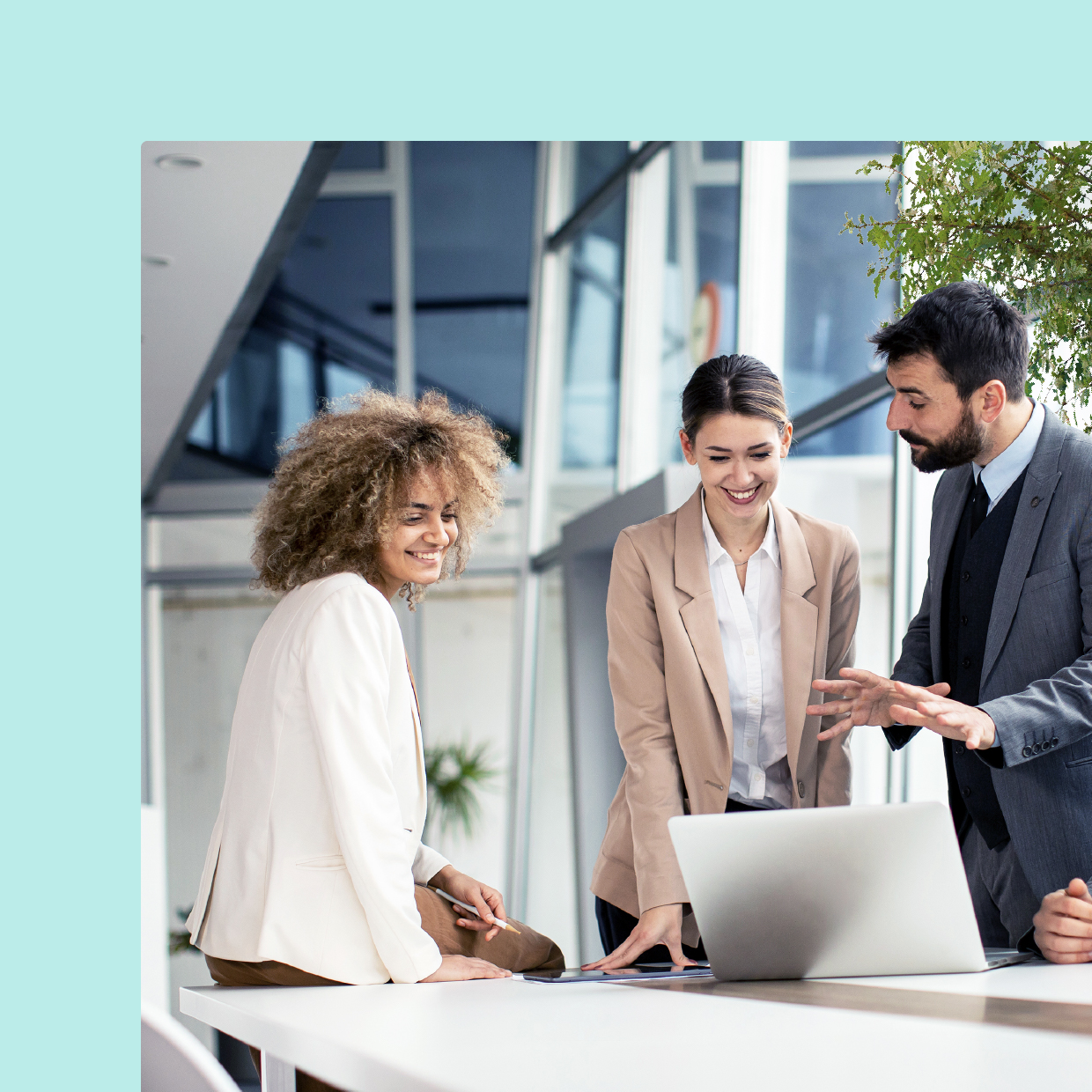 Three business professionals, one woman with curly hair and two in blazers, interacting and discussing over a laptop at a bright workspace with plants in the background.