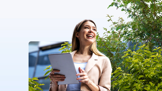 Young woman standing outdoors holding a tablet with a smile, green trees in the background.